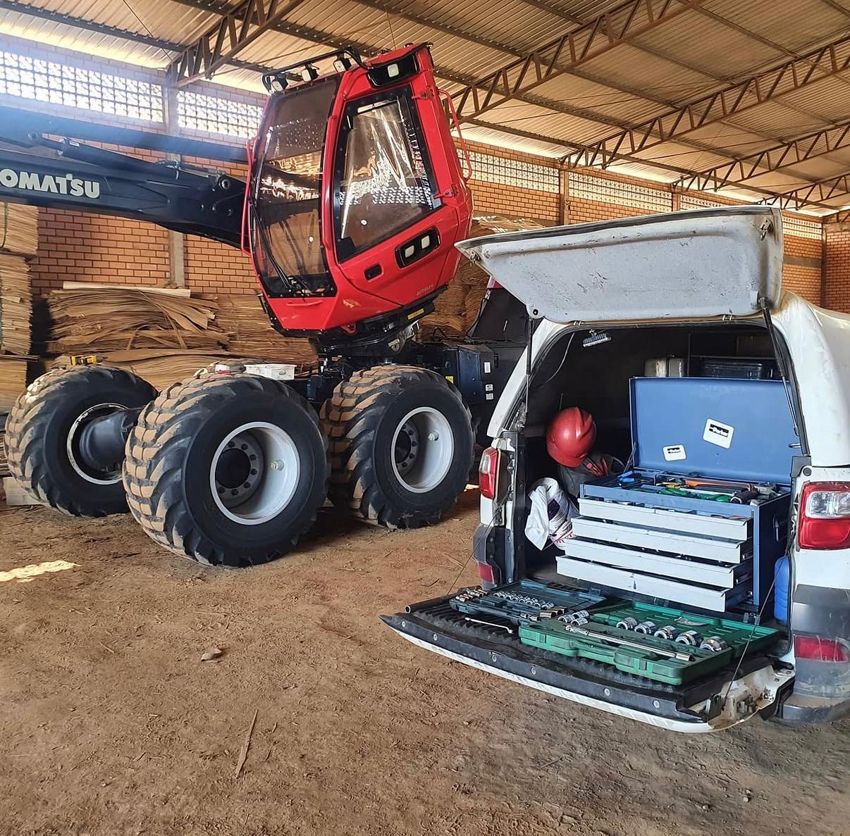 An organized outdoor service area dedicated to forest machinery maintenance, featuring a stationary harvester and forwarder parked on a clean gravel pad. In the foreground, a selection of new, clean components—hydraulic cylinders, steel pins, hoses, bearings, and reinforced grapples—is neatly arranged on sturdy pallets and metal racks. The machines stand behind them as a clear demonstration of compatibility and support. Late afternoon natural light casts long, soft shadows, bringing out metallic reflections and surface details. Shot from a slightly elevated, wide-angle perspective in sharp focus, the photographic composition communicates professionalism, readiness, and technical expertise, with a calm, orderly atmosphere and no people or branding visible.